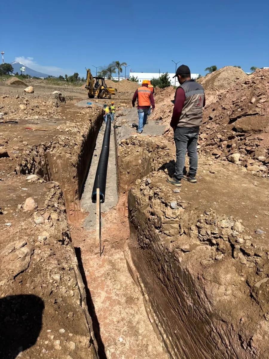 Tubería corrugada negra tendida en zanja larga, con retroexcavadora, personal y el volcán al fondo